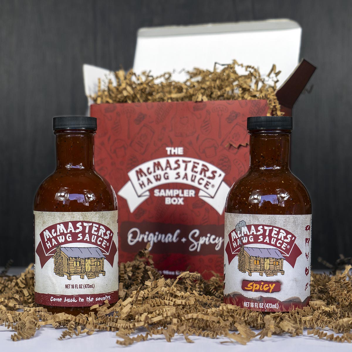 A bottle of McMasters' Hawg Sauce Original and a bottle of Spicy in front of a red gift box labeled The McMasters' Hawg Sauce Sampler Box